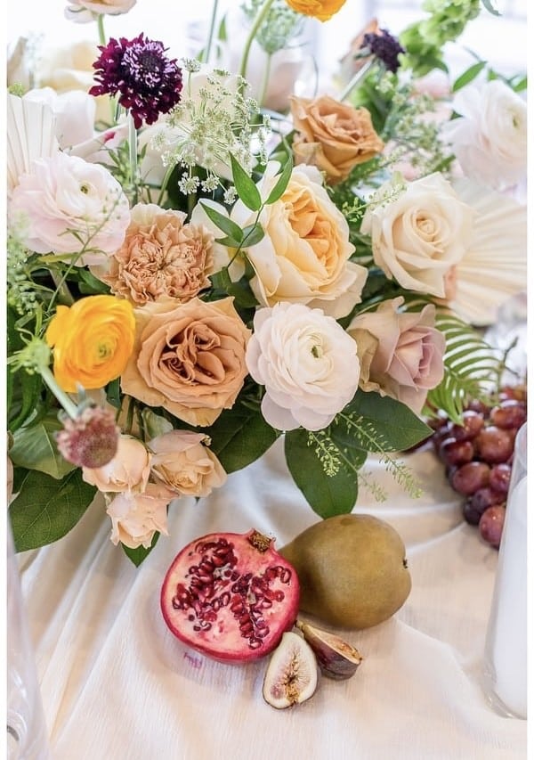 A floral arrangement with roses and assorted flowers sits on a table next to a halved pomegranate, a fig, a pear, and a small bunch of grapes—perfect for an elegant Wequassett Christmas Dinner setting.