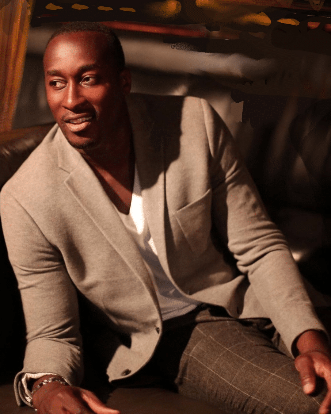 A man wearing a light gray blazer and white shirt sits on a dark couch, looking to the side and smiling in a warmly lit setting at the Cape Cod Arts Festival.