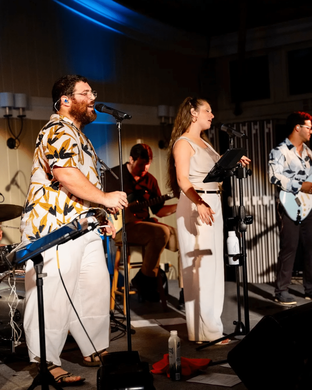 A band performs on stage at the Cape Cod Jazz Festival, with two vocalists at the front, accompanied by musicians playing guitar and drums under warm lighting.