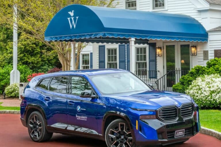 A blue BMW SUV is parked in front of a building with a blue awning displaying a white “W” and a bird logo, hinting at Wequassett Resort. The car has “BMW” and “BMW Security” decals on its doors.
