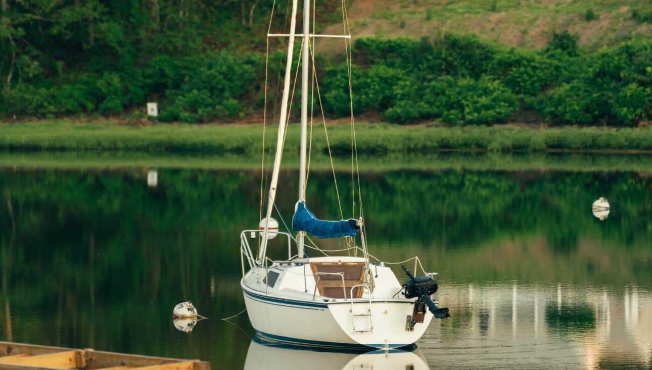 A small white sailboat with a covered sail is anchored on calm water near the Wequassett Resort, surrounded by lush greenery along the riverbank in the background.