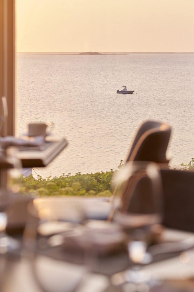 A small motorboat floats on calm water at sunset, viewed from a restaurant table set with glasses and plates in the foreground.
