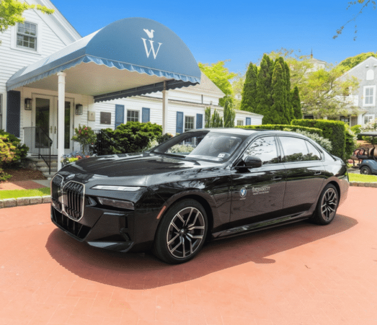 A black BMW sedan is parked on a red brick driveway in front of a white building with blue shutters and a canopy labeled with a "W," evoking the classic elegance of Wequassett Resort & Golf Club.