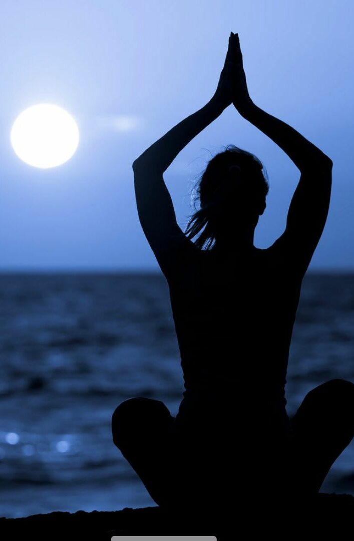 A person sits cross-legged on the beach, silhouetted against the ocean, with hands raised above their head at sunset—capturing the serenity of a Women's Wellness Retreat.