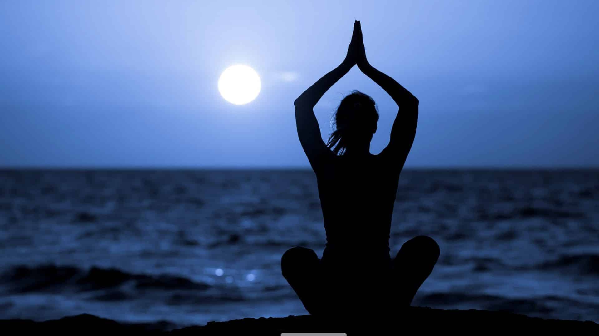 A person sits cross-legged on the beach, silhouetted against the ocean, with hands raised above their head at sunset—capturing the serenity of a Women's Wellness Retreat.