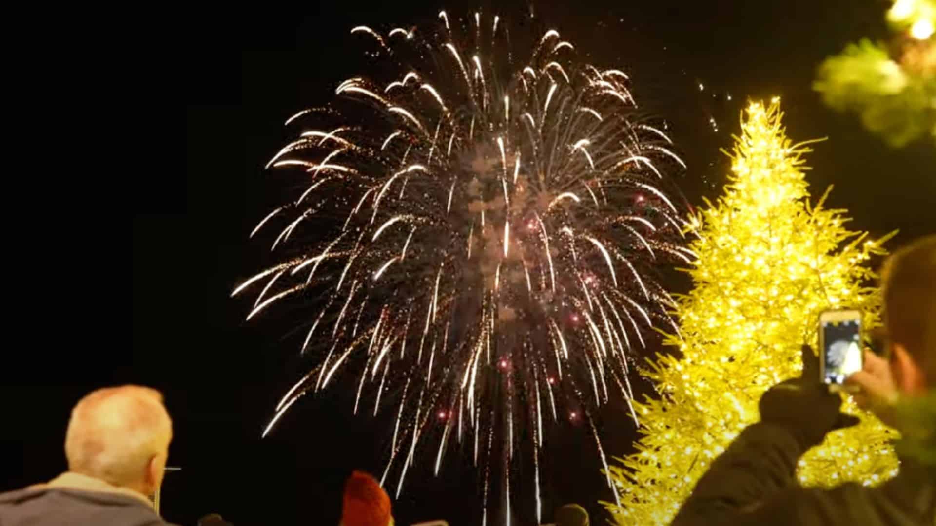 People watch a fireworks display at night near a brightly lit Christmas tree during the Merry Market; one person is capturing the Tree Lighting scene on a smartphone.