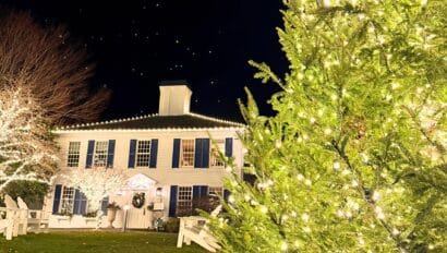 A large Christmas tree decorated with lights stands in front of a white colonial-style house adorned with holiday lights at night, creating a festive celebration and tree lighting scene.