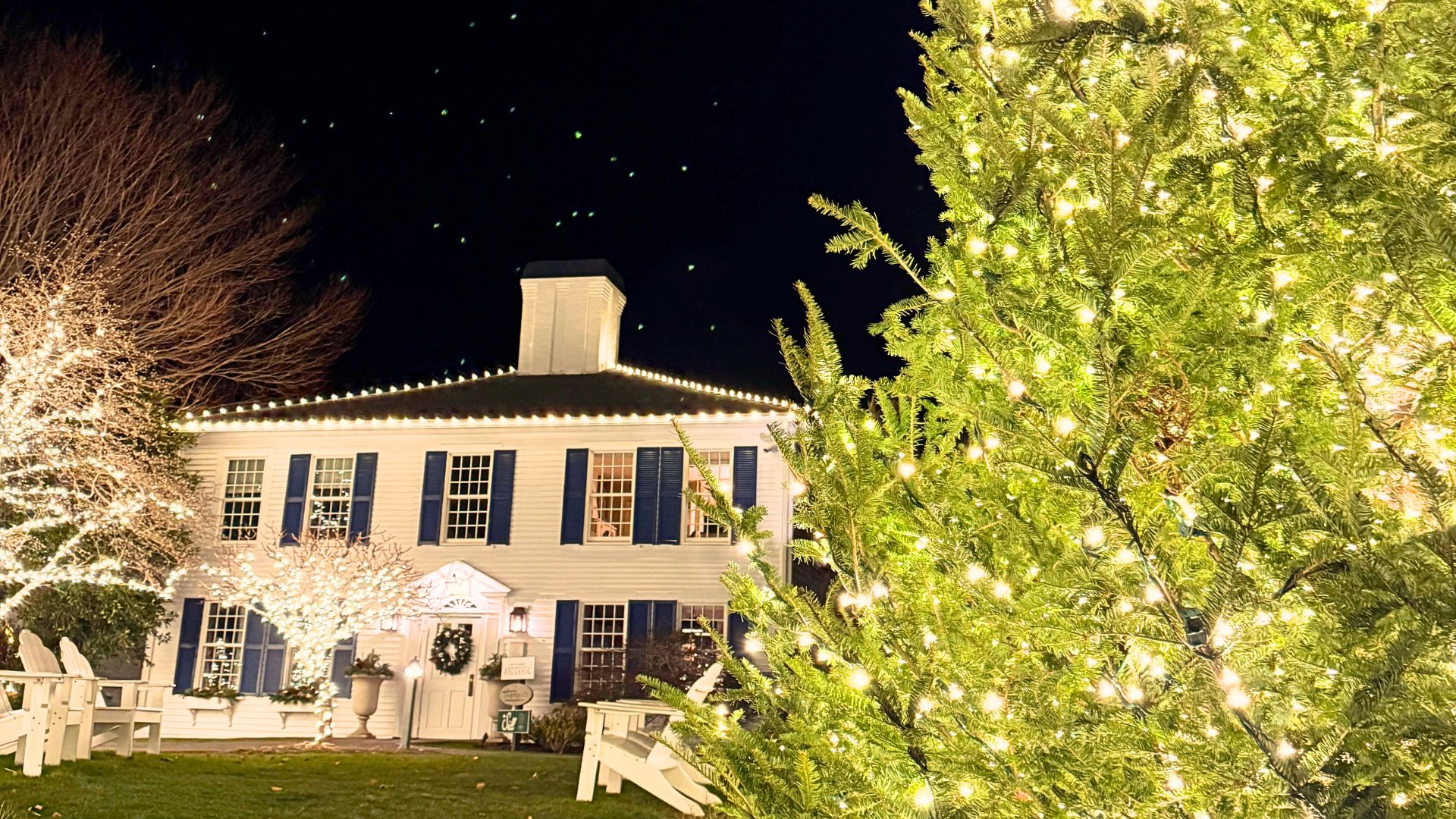 A large Christmas tree decorated with lights stands in front of a white colonial-style house adorned with holiday lights at night, creating a festive celebration and tree lighting scene.