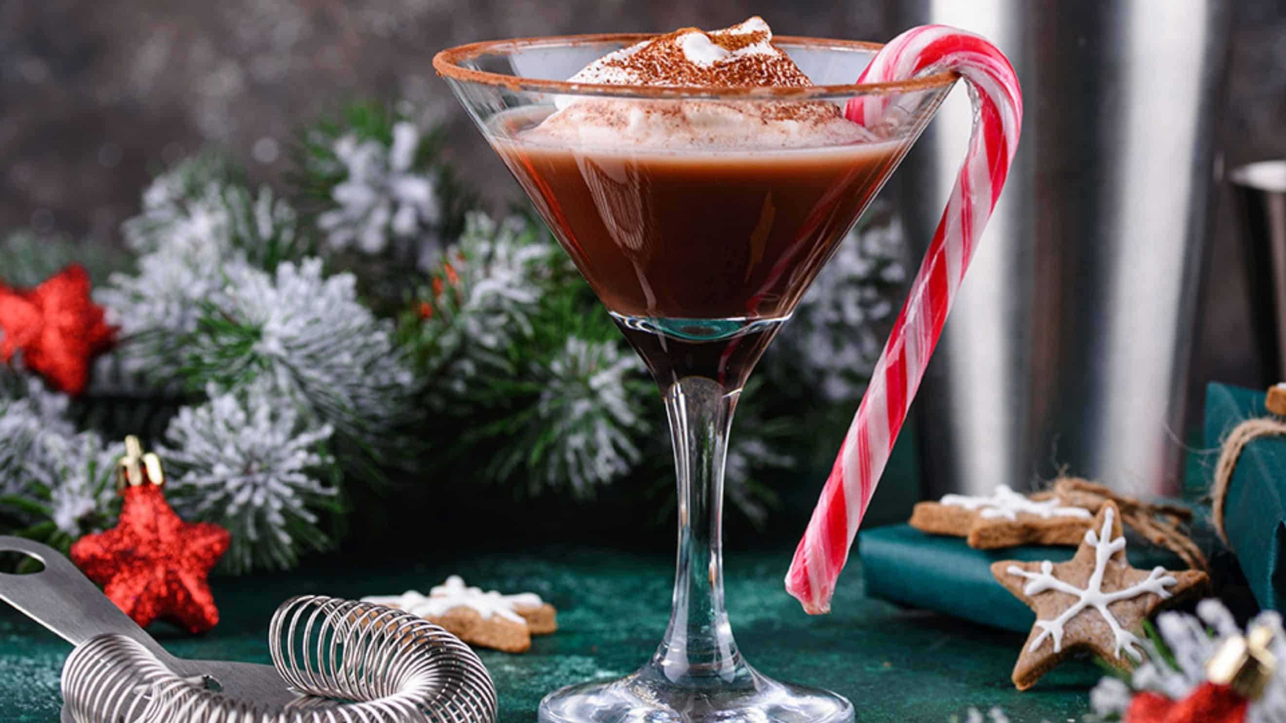 A martini glass filled with a Naughty & Nice chocolate cocktail, topped with whipped cream and cocoa powder, garnished with a candy cane. Holiday decorations and gingerbread cookies set the perfect festive mixology scene in the background.