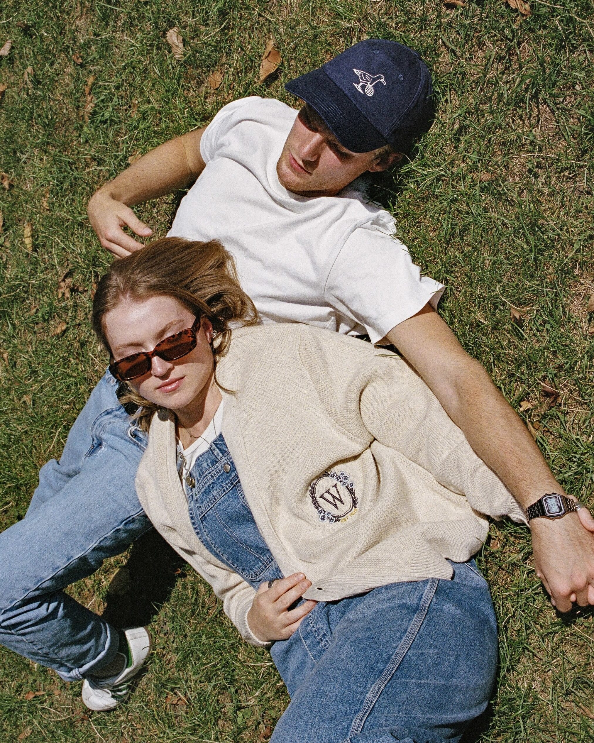 Two people lie on grass, enjoying a sunny day. The woman wears sunglasses, a cream jacket, and jeans; the man sports a navy cap, white shirt, and jeans. Their relaxed vibe suggests effortless collaboration outdoors.