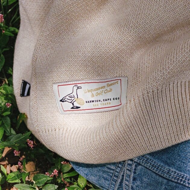 A beige sweater with a "Wiquasasset Resort & Golf Club" patch is shown, partially covering denim and surrounded by green leaves and small pink flowers—perfect for a cozy evening or stylish Dinner Service NY.