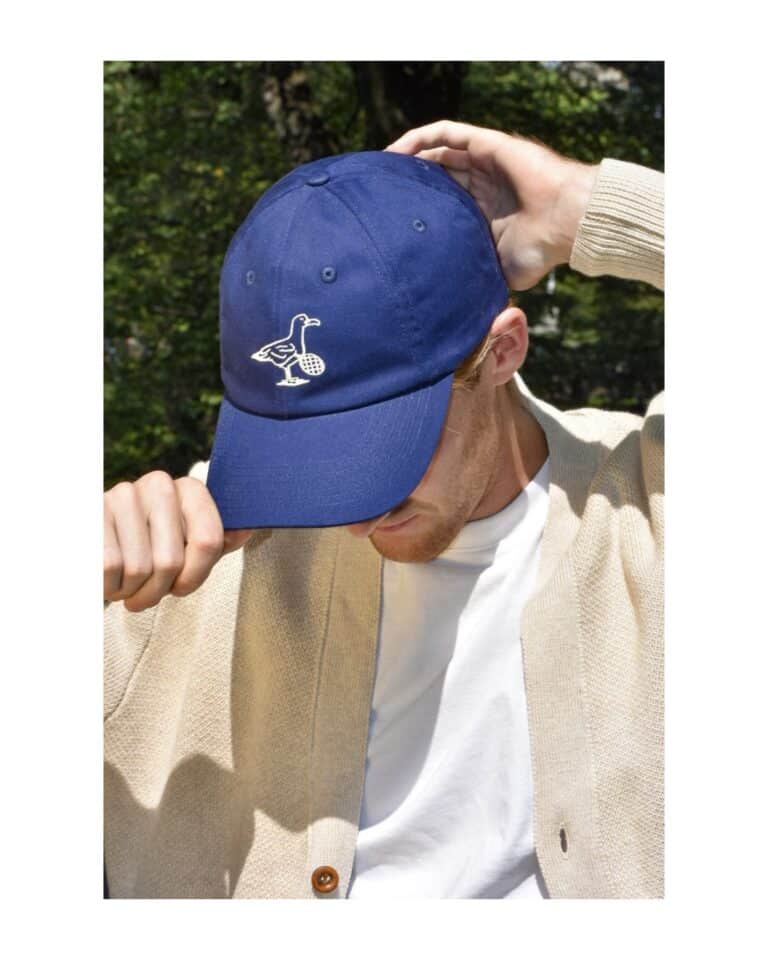 A person wearing a blue cap with a white goose and soccer ball design, a beige cardigan, and a white shirt, holding the brim of the cap outdoors after enjoying Dinner Service NY.