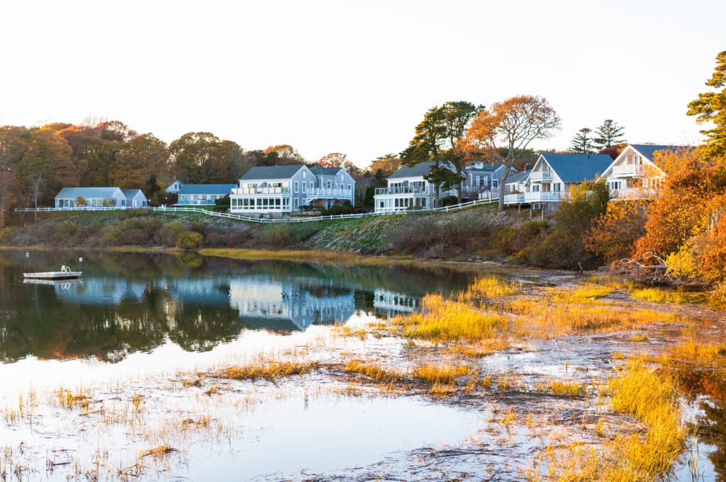 Waterfront houses are reflected in a calm body of water, surrounded by autumn trees and grasses, evoking the vibrant spirit of Fall Festivals, with a small boat visible on the left side of the image.