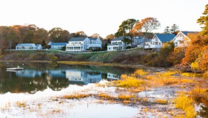 Waterfront houses are reflected in a calm body of water, surrounded by autumn trees and grasses, evoking the vibrant spirit of Fall Festivals, with a small boat visible on the left side of the image.