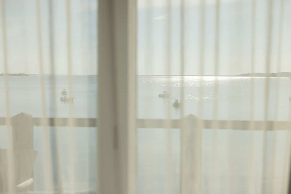 Sheer curtains partially obscure a view of calm water with several small boats and a wooden railing, under a bright sky.