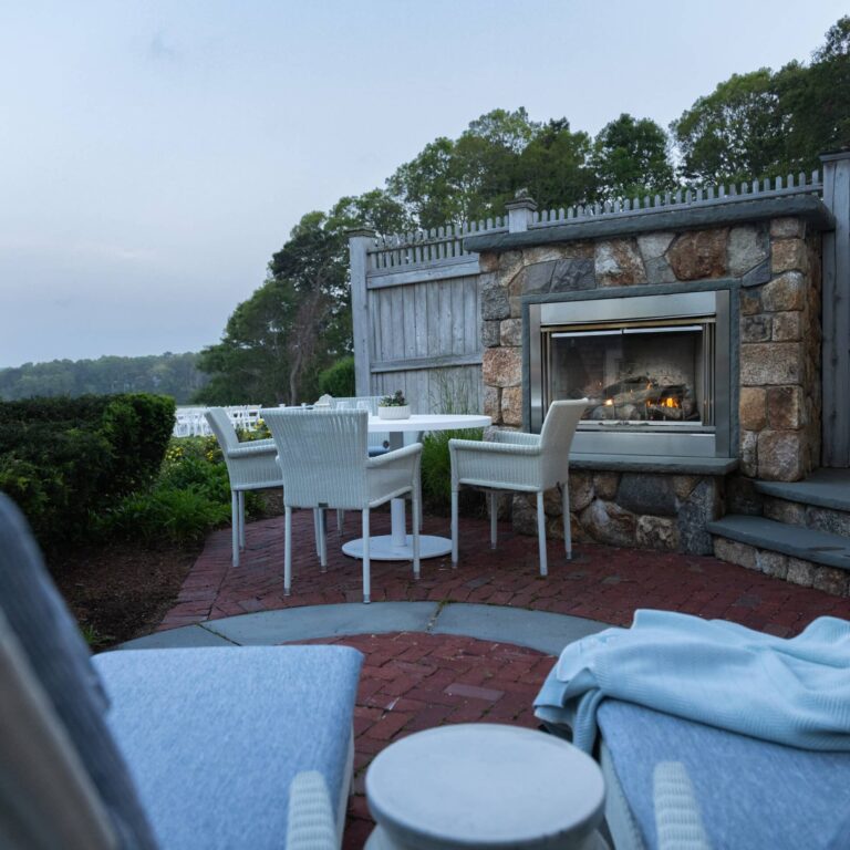 Outdoor patio at Wequassett Resort and Golf Club with white wicker chairs around a stone fireplace, set on a red brick floor beside greenery and a wooden fence, with blue-cushioned lounge chairs—an inviting Cape Cod wedding venue.