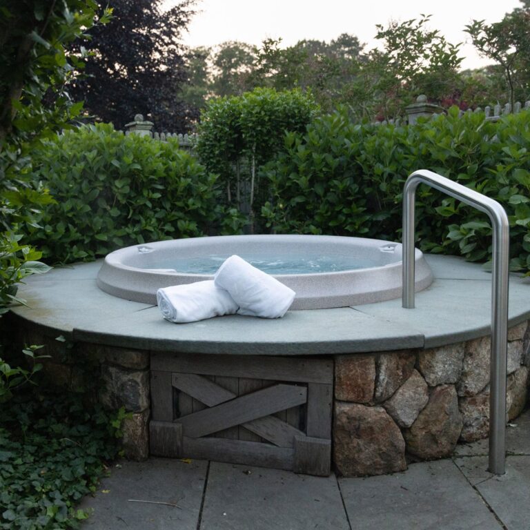 Round outdoor hot tub surrounded by greenery at a Cape Cod hotel, with two rolled white towels on the stone edge and a metal handrail on the right—perfect relaxation after a day at the nearby Cape Cod golf course.