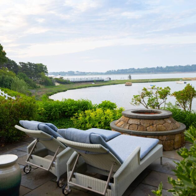 Two cushioned lounge chairs face a stone fire pit on a patio at this Cape Cod hotel, overlooking a river with boats and lush greenery under a cloudy sky.