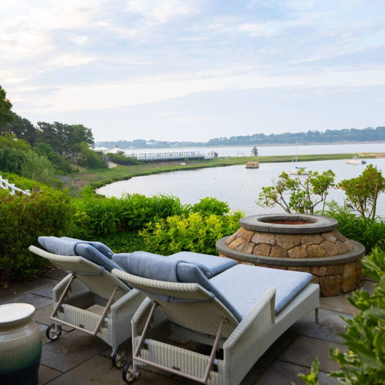 Two cushioned lounge chairs face a stone fire pit on a patio at this Cape Cod hotel, overlooking a river with boats and lush greenery under a cloudy sky.