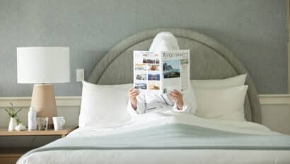 A person in a white robe and towel sits in bed reading a magazine, with a lamp, plant, and cups on the bedside table—perhaps browsing for special offers.