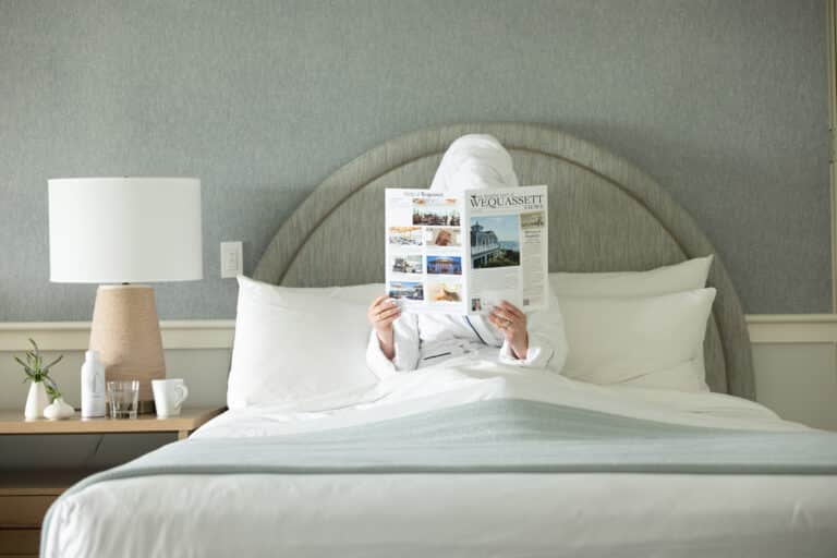 A person in a white robe and towel sits in bed reading a magazine, with a lamp, plant, and cups on the bedside table—perhaps browsing for special offers.