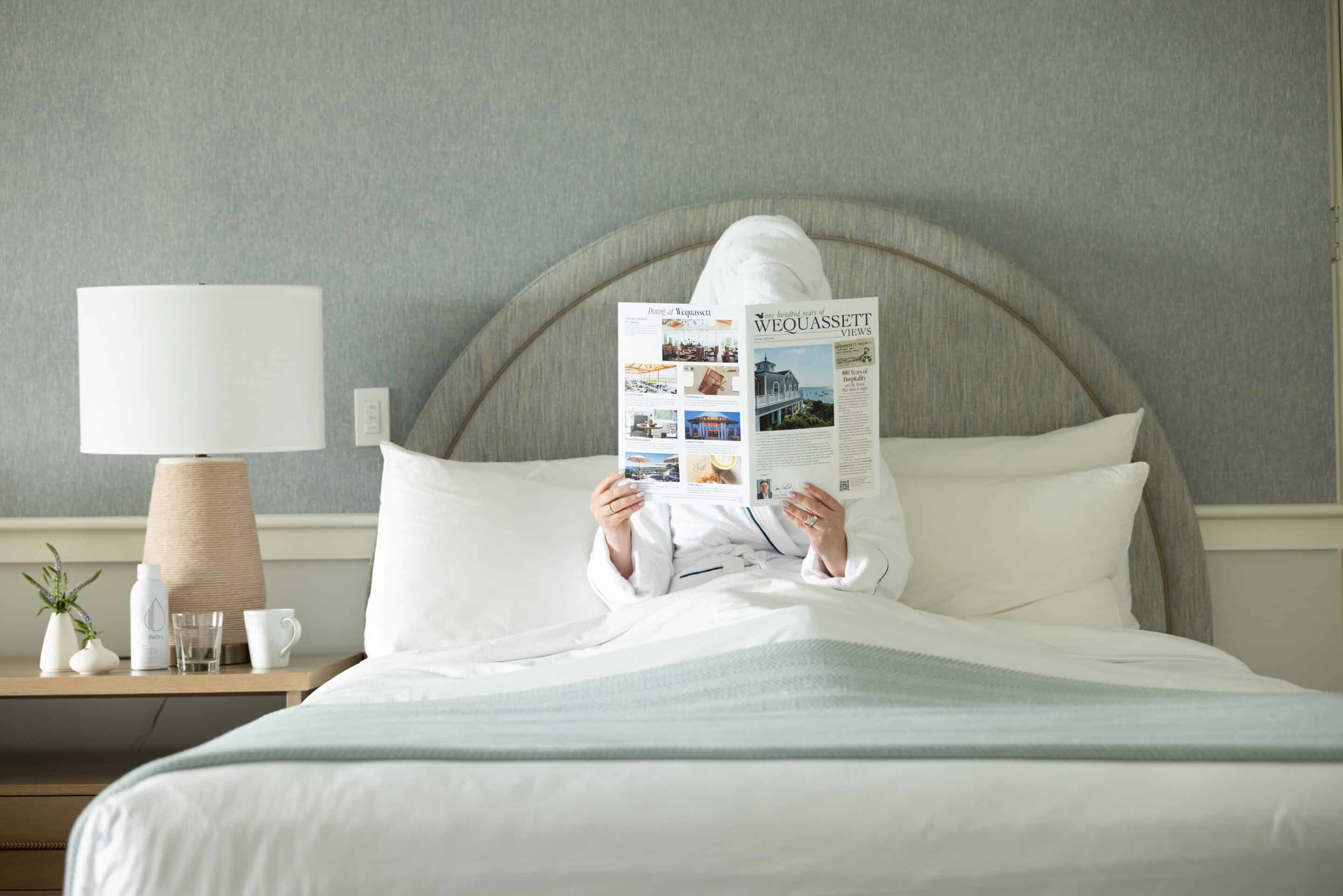 A person in a white robe and towel sits in bed reading a magazine, with a lamp, plant, and cups on the bedside table—perhaps browsing for special offers.