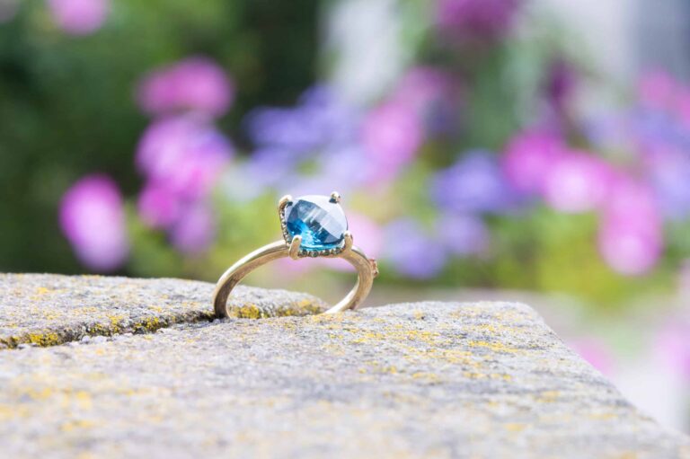 A gold ring with a blue gemstone is placed on a stone surface, celebrating Wequassett’s centennial amid blurred green foliage and purple flowers.