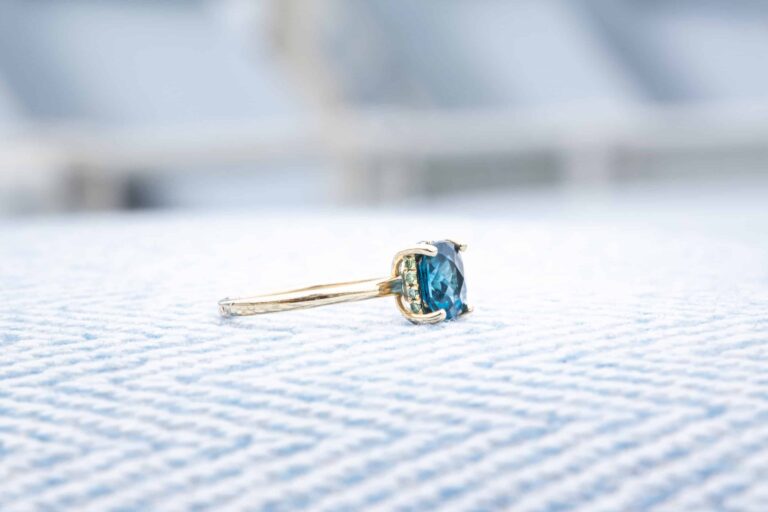 A gold ring with a blue gemstone rests on a light blue, textured surface, echoing the timeless elegance of Wequassett Resort as it celebrates 100 years, set against a beautifully blurred background.