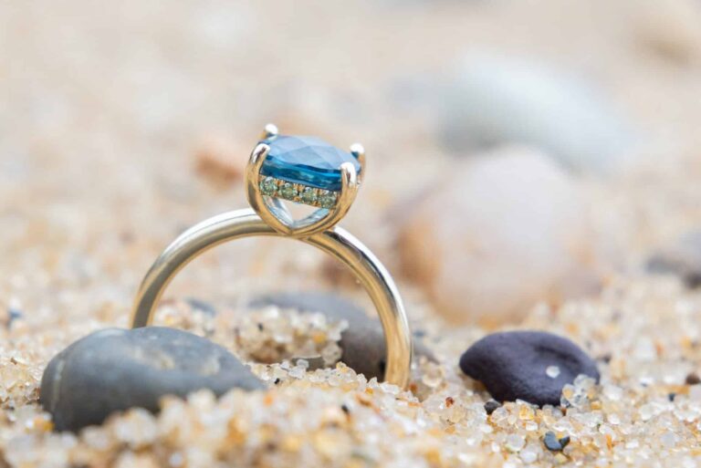A gold ring with a blue gemstone stands upright in sandy ground, surrounded by small stones and pebbles—a timeless treasure fitting to celebrate Wequassett’s centennial.