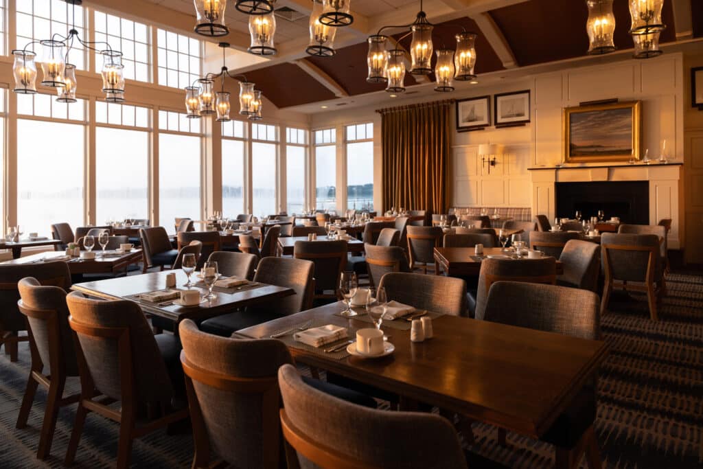 Spacious dining room with neatly set tables and cushioned chairs, featuring large windows with natural light and chandeliers hanging from a high ceiling at Wequassett Resort.