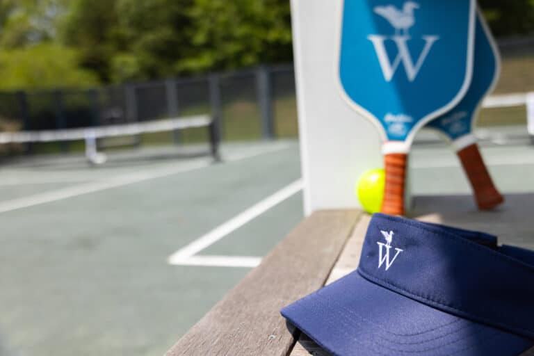 A navy visor and two pickleball paddles, essential gear for racquet sports enthusiasts, rest on a bench near the pickleball court, with a yellow ball on the ground.