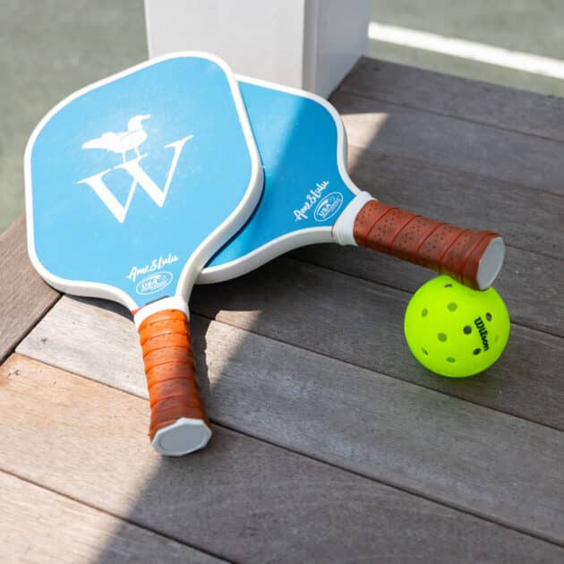 Two blue pickleball paddles with brown handles and a yellow pickleball rest on a wooden surface next to a court—perfect for fans of racquet sports.
