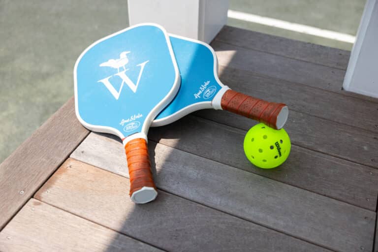 Two blue pickleball paddles with brown handles and a yellow pickleball rest on a wooden surface next to a court—perfect for fans of racquet sports.