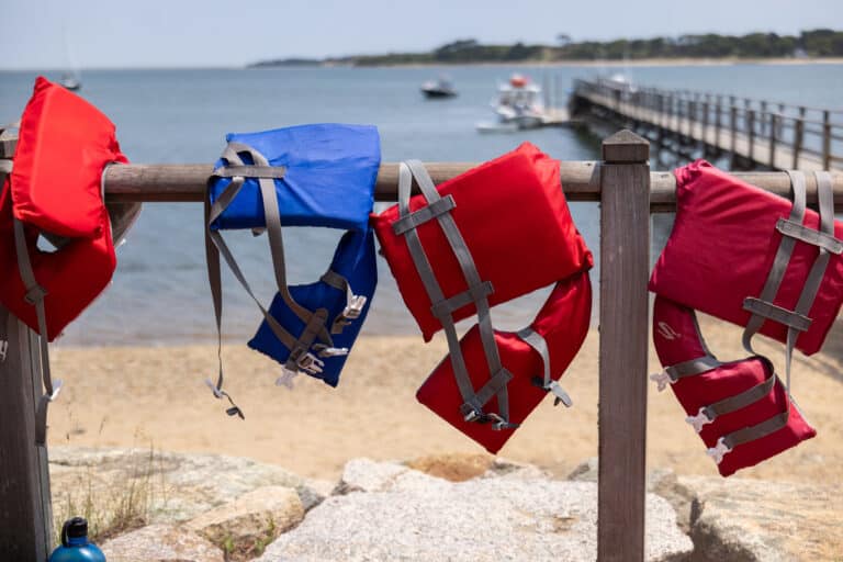 Four red and one blue life jackets hang on a wooden railing near a sandy beach at Pleasant Bay, with a pier, boats, and the floating classroom’s solar-powered vessel visible in the background.