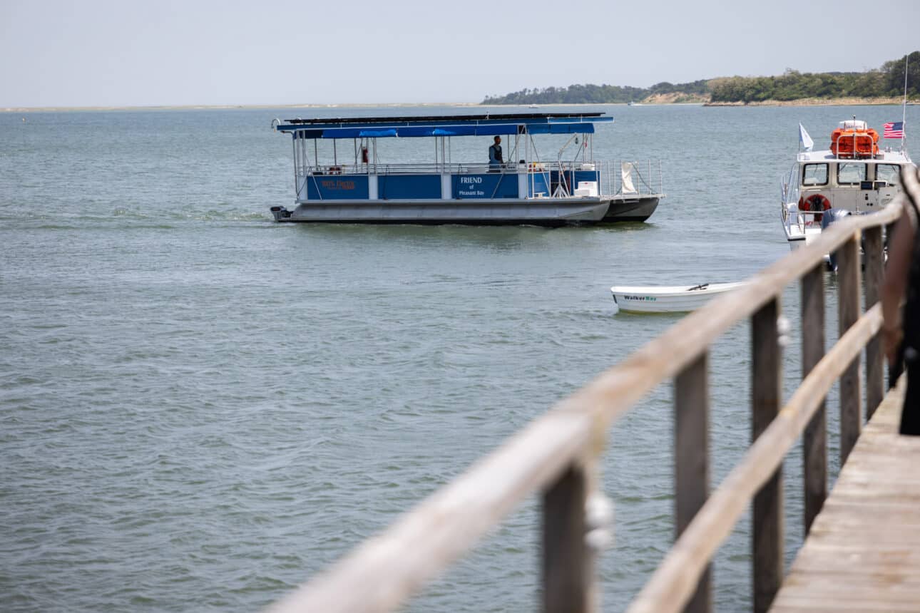 A blue and white ferry boat, known as The Friend, moves across the calm waters of Pleasant Bay, with a dock and a smaller boat in the foreground. Trees line the distant shoreline.