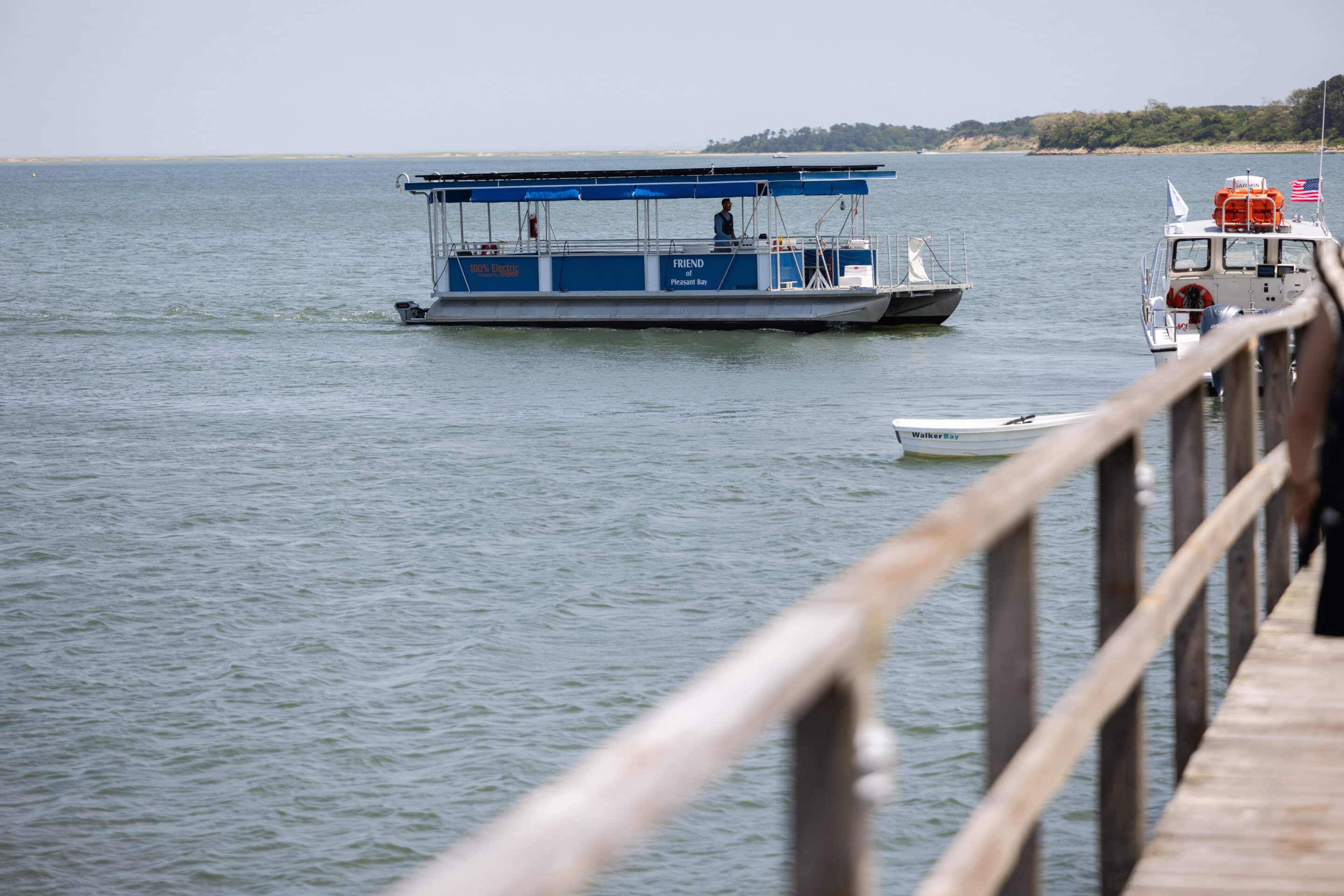 A blue and white ferry boat, known as The Friend, moves across the calm waters of Pleasant Bay, with a dock and a smaller boat in the foreground. Trees line the distant shoreline.