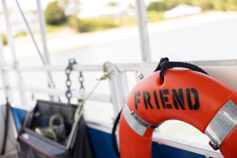 An orange life preserver labeled "FRIEND" hangs on the railing of a solar-powered boat, with ropes and chains visible nearby as it cruises Pleasant Bay.