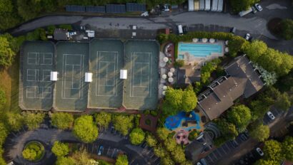 Aerial view of a sports facility featuring five racquet sports tennis courts, a swimming pool, a playground, surrounding buildings, and parking areas bordered by trees.