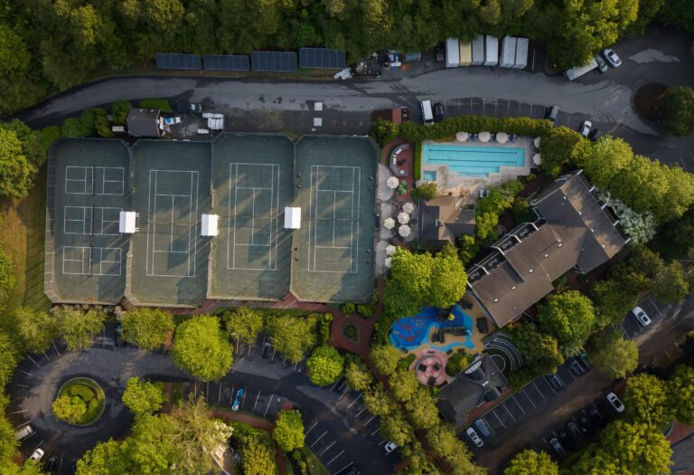 Aerial view of a sports facility featuring five racquet sports tennis courts, a swimming pool, a playground, surrounding buildings, and parking areas bordered by trees.