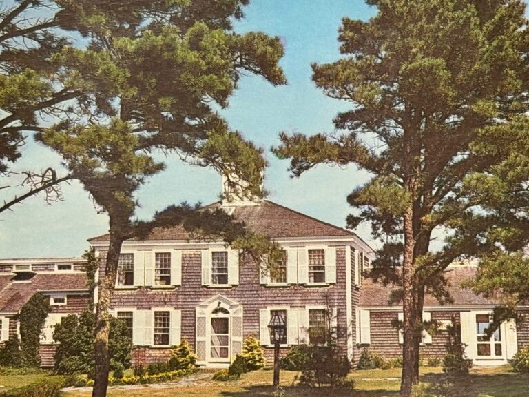 A two-story house with wood shingles and white trim, surrounded by trees and landscaping, is shown on a sunny day—capturing the timeless charm of Wequassett as it celebrates its 100 Years anniversary.