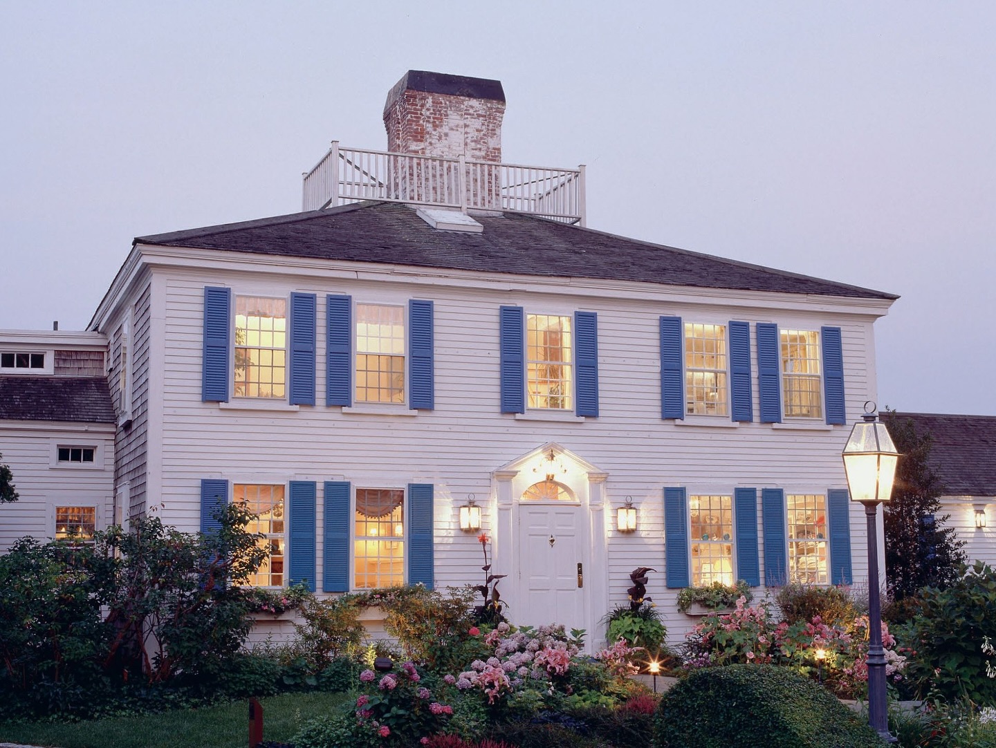 A two-story white house with blue shutters, a central chimney, and a garden in front, photographed at dusk with lights on inside, reminiscent of Wequassett’s charm as it celebrates 100 years of timeless beauty.