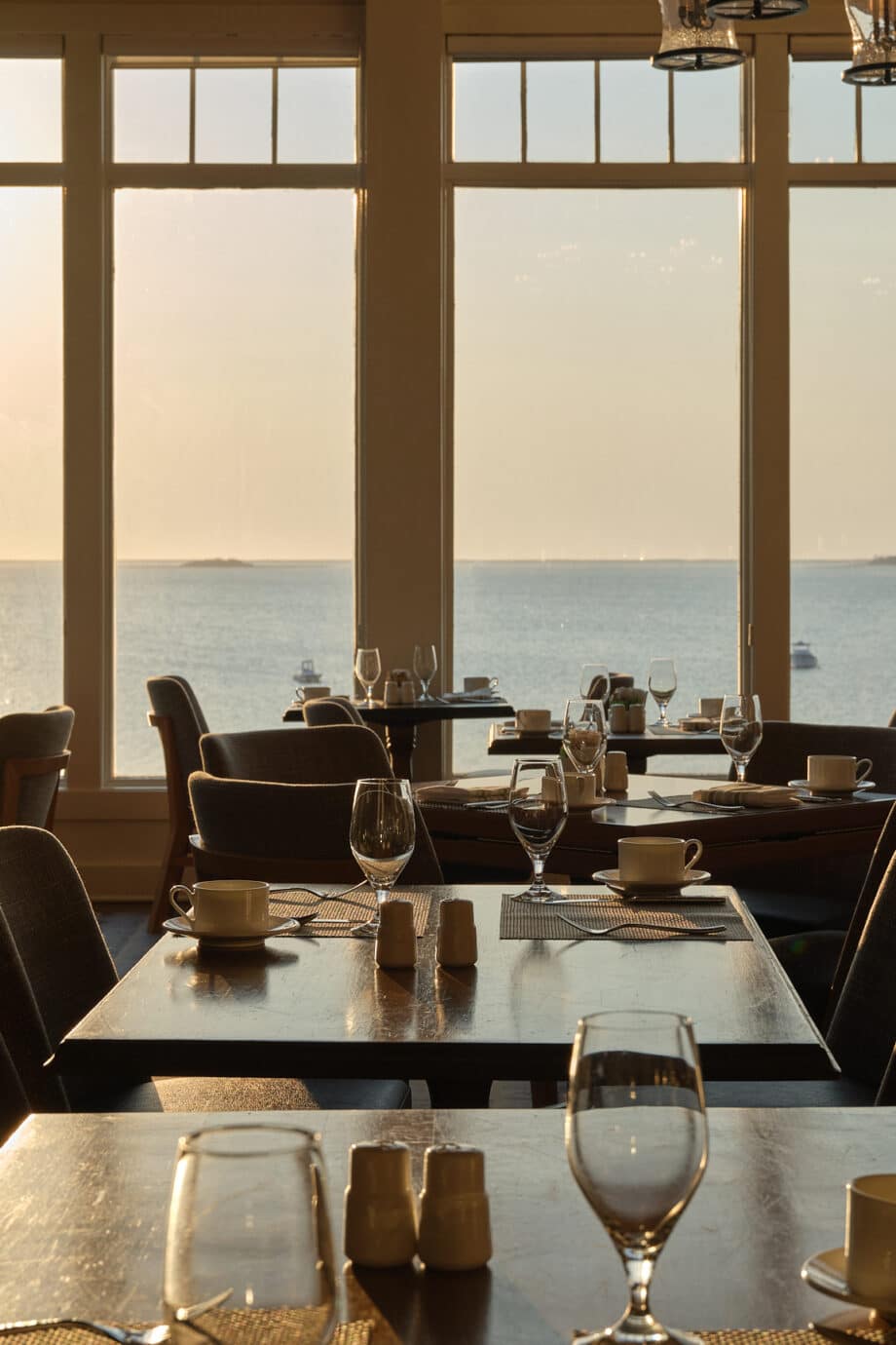 Sunlit restaurant dining area with set tables and empty chairs overlooks a calm body of water through large windows, perfect for a serene New Year’s Day brunch at sunset.