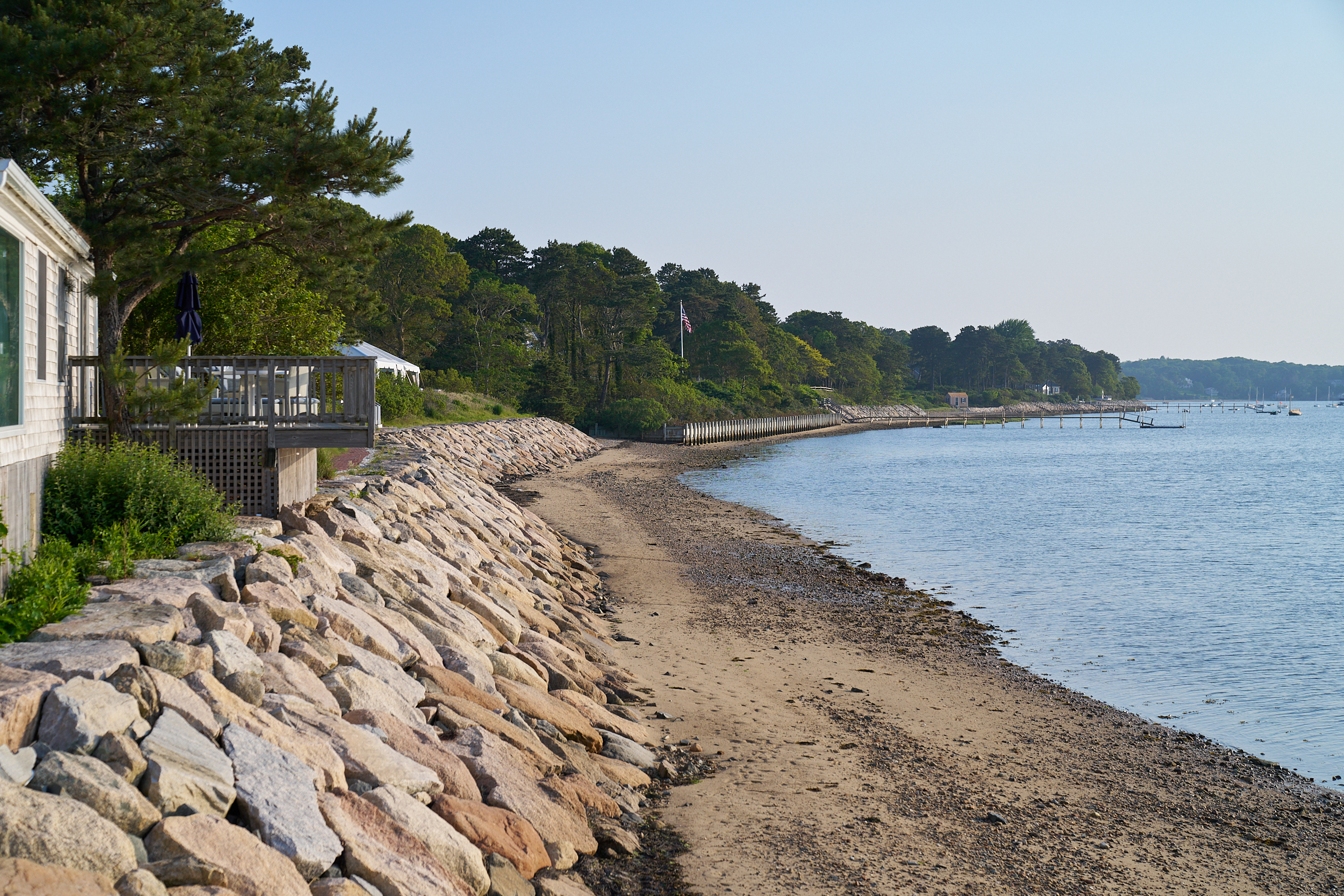 House with a deck overlooks a rocky shoreline and sandy beach with calm water, trees in the distance, and clear sky—a perfect retreat that embraces sustainability in its design and setting.