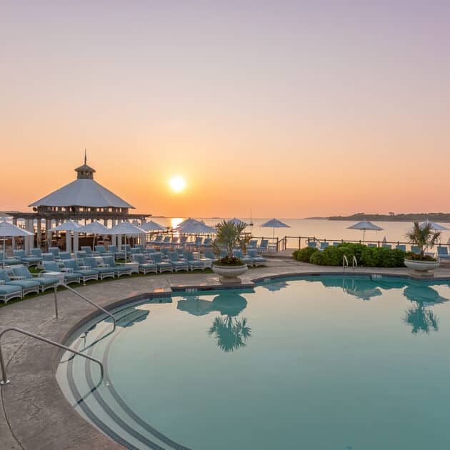 A round outdoor pool with lounge chairs, umbrellas, and a gazebo overlooks a calm body of water at sunrise or sunset.