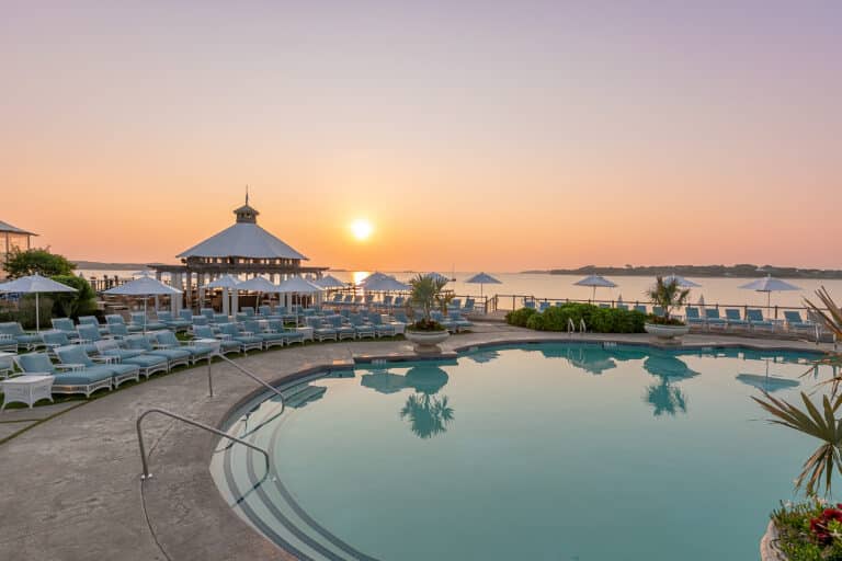 A round outdoor pool with lounge chairs, umbrellas, and a gazebo overlooks a calm body of water at sunrise or sunset.
