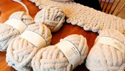 A person arm-knits a chunky, beige blanket on a wooden table with four large skeins of matching yarn nearby, inspired by cozy moments at Wequassett Resort and Golf Club—a premier Cape Cod wedding venue.