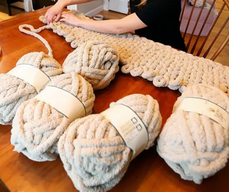 A person arm-knits a chunky, beige blanket on a wooden table with four large skeins of matching yarn nearby, inspired by cozy moments at Wequassett Resort and Golf Club—a premier Cape Cod wedding venue.