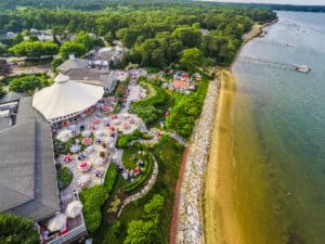 Aerial view of a waterfront venue with outdoor seating, red and white tablecloths, gardens, buildings, and a rocky shoreline next to calm water.