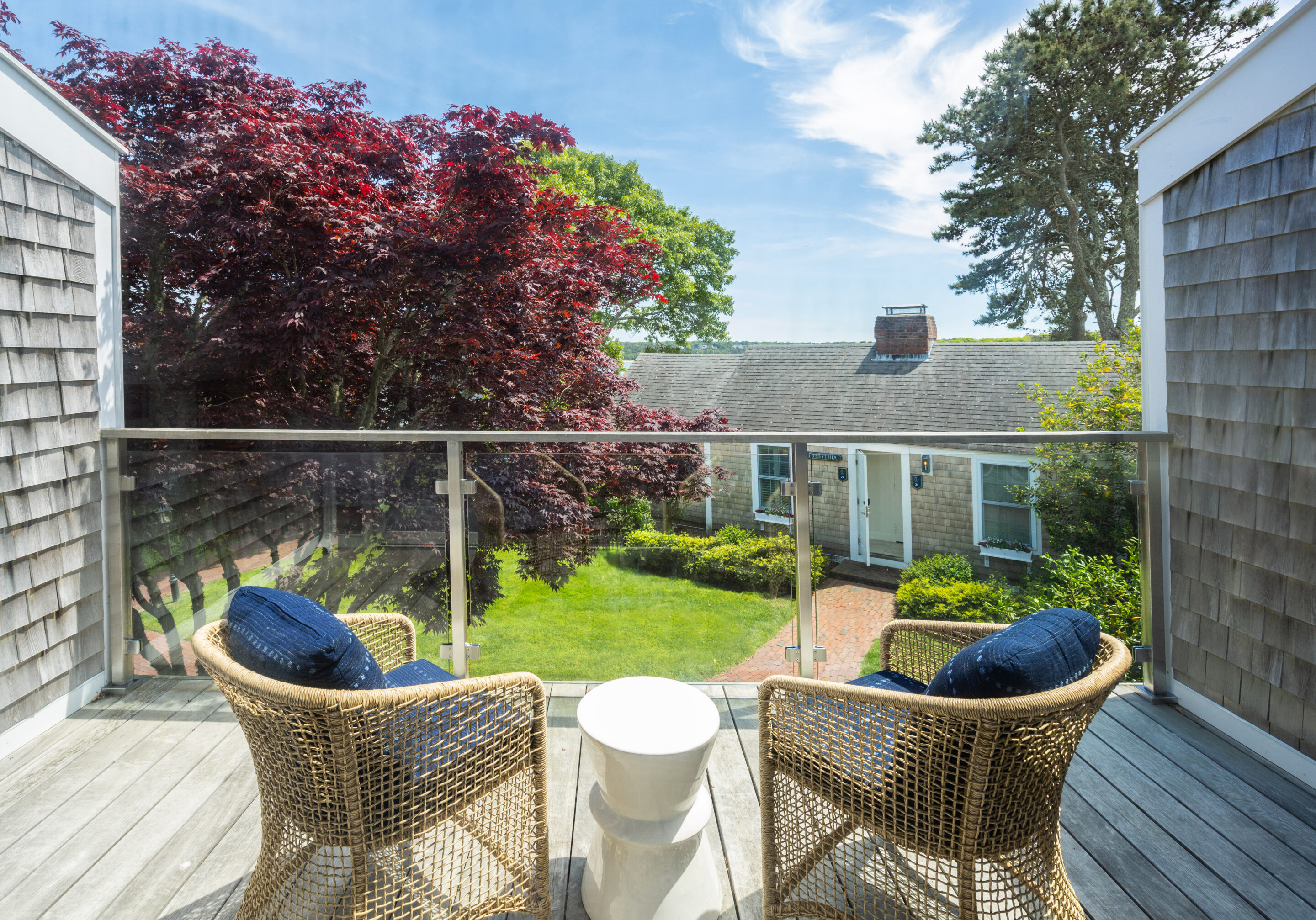 Two wicker chairs with navy cushions face a glass railing on a deck, overlooking a green yard dotted with sweet pea blooms, a red tree, and a house with gray shingles on a sunny day.