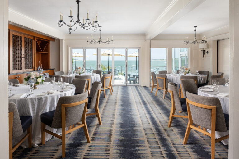 A bright private dining room features round tables set for a meal, upholstered chairs, chandeliers, floral centerpieces, and large windows overlooking an outdoor patio and water.